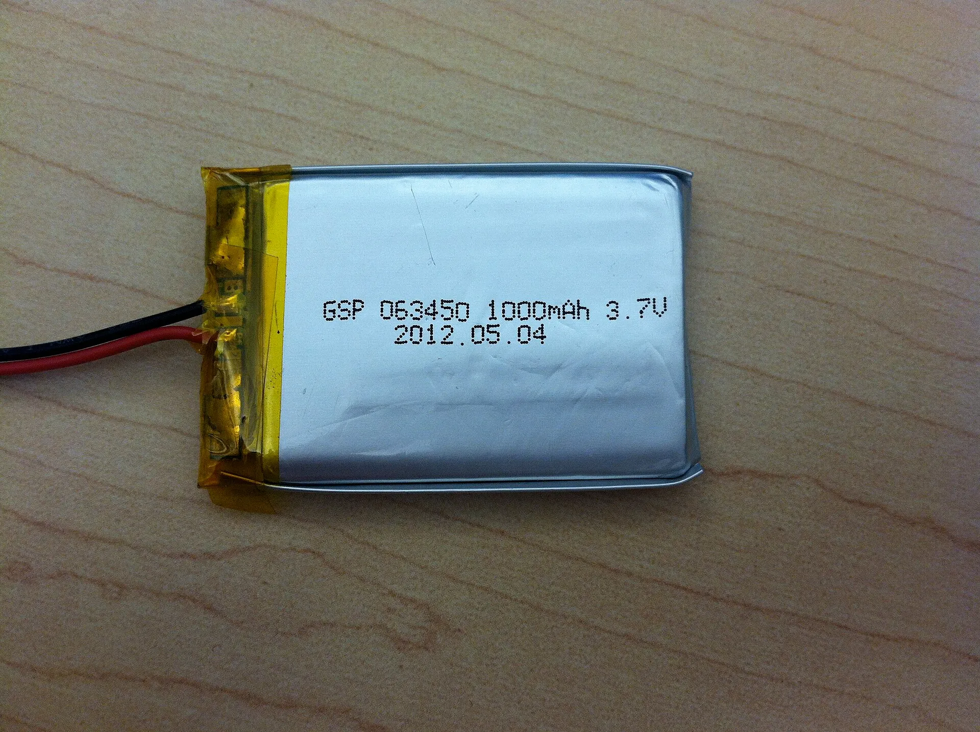 Batteria piatta e rettangolare in un involucro argentato, appoggiata su un tavolo di legno, con la scritta 1000 mAh 3.7V e due fili, uno rosso e uno nero, che escono da un lato protetto da nastro giallo.