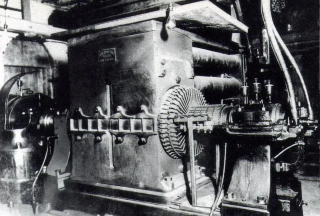 Fotografia d'epoca in bianco e nero di una grande dinamo in una sala macchine: un massiccio blocco metallico con un anello di spazzole a raggiera e cavi che scendono dall'alto.