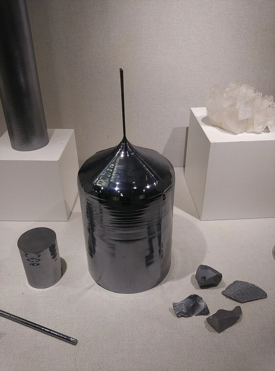 Un lingotto cilindrico di silicio grigio scuro e lucido, con la sommità a cono che termina in un sottile collo verticale, esposto in una vetrina accanto a un cilindro più piccolo, a frammenti irregolari di silicio grezzo e, su un piedistallo, a un cristallo di quarzo bianco.