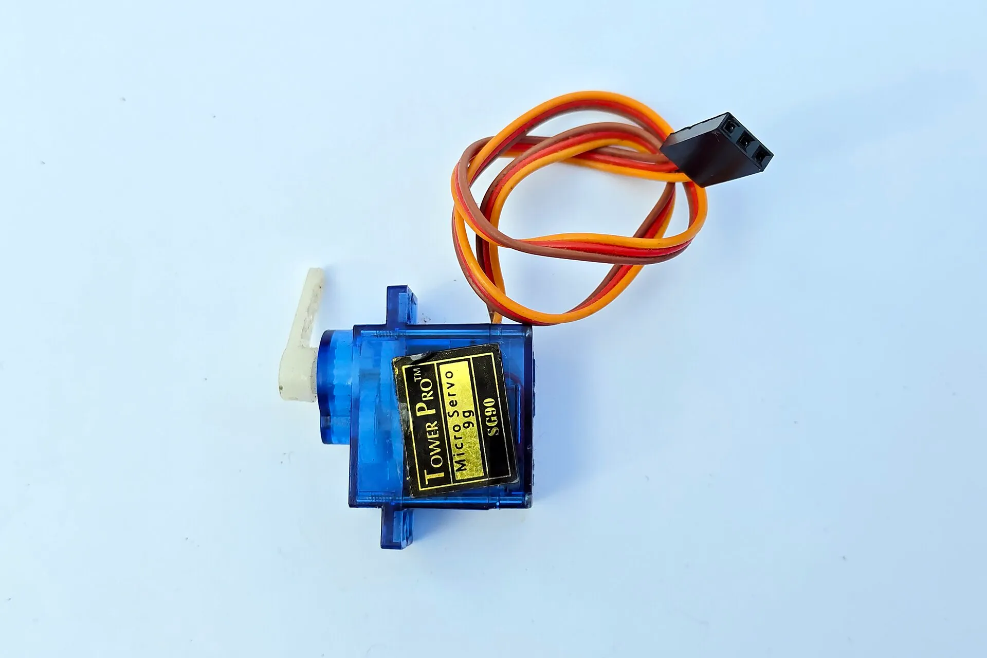 Micro servomotore in plastica blu trasparente con l'etichetta Tower Pro Micro Servo 9g SG90, una squadretta bianca sull'albero e un cavo a tre fili, marrone, rosso e arancione, con un connettore nero.