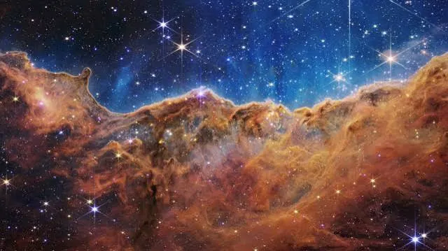 Nubi di gas e polvere dai toni arancio che si alzano come una parete di roccia sotto un cielo blu pieno di stelle.